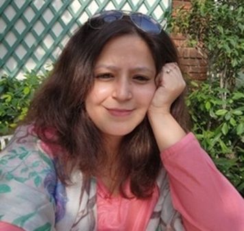 Portrait of a woman with shoulder-length brown hair and a pink top, sunglasses on her head, resting her head on her hand in a garden.