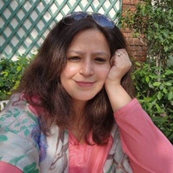 Portrait of a woman with shoulder-length brown hair and a pink top, sunglasses on her head, resting her head on her hand in a garden.