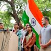 Group of people at a press conference outdoors, Indian flag in the background. microphones are on the podium.