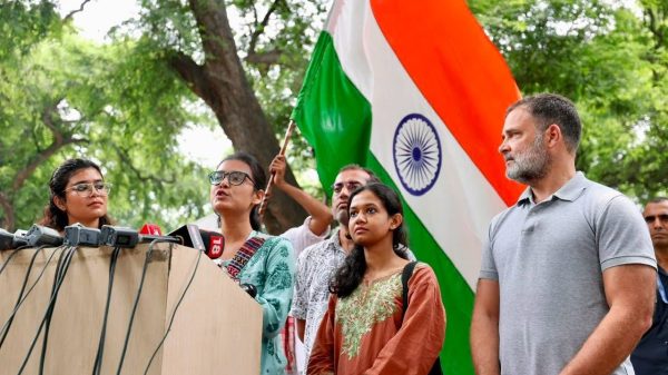Group of people at a press conference outdoors, Indian flag in the background. microphones are on the podium.