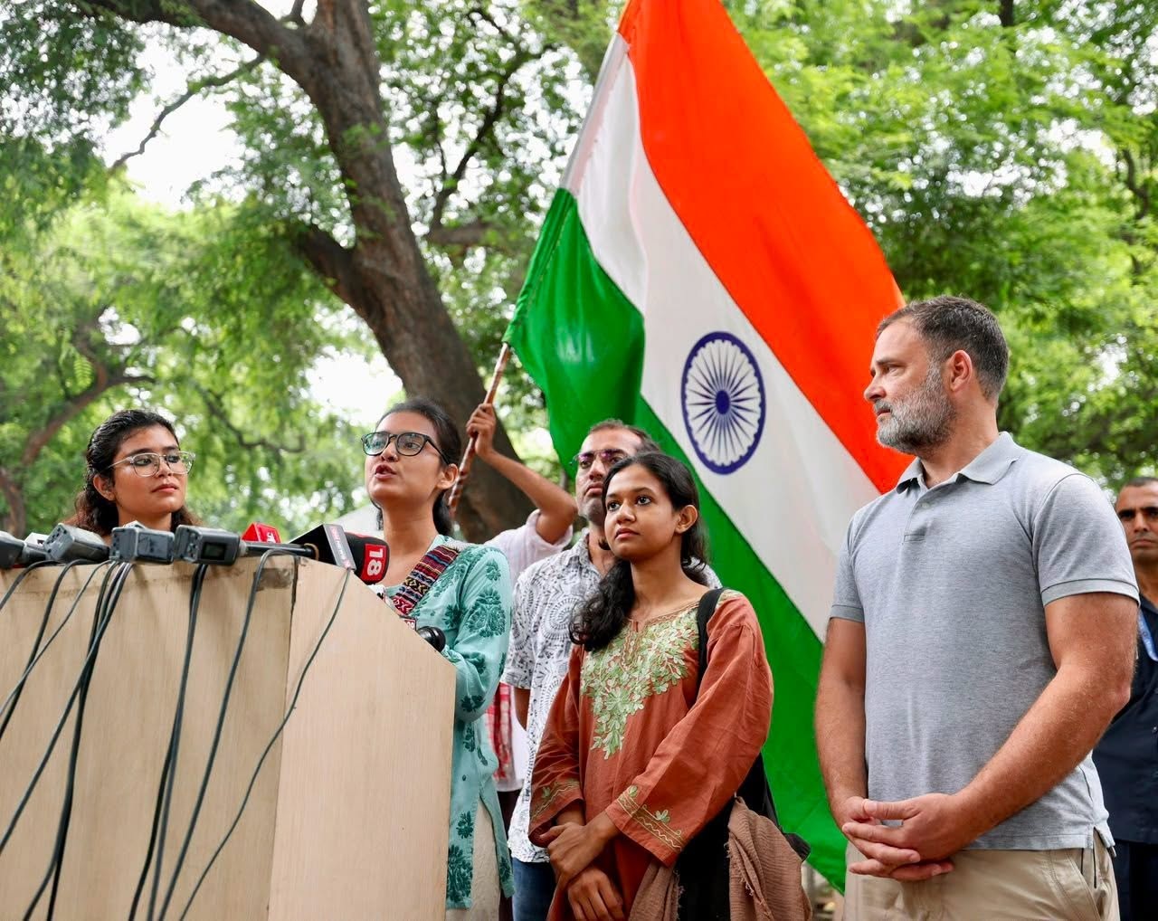 Group of people at a press conference outdoors, Indian flag in the background. microphones are on the podium.
