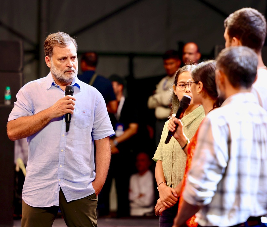 rahul gandhi | No. 1 Indian Media News Portal Man with a microphone in a light blue shirt speaks on stage, while a small group listens in the foreground and background.