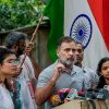 Man with gray beard addresses a crowd at a press conference, microphones in front, Indian flag in the background.