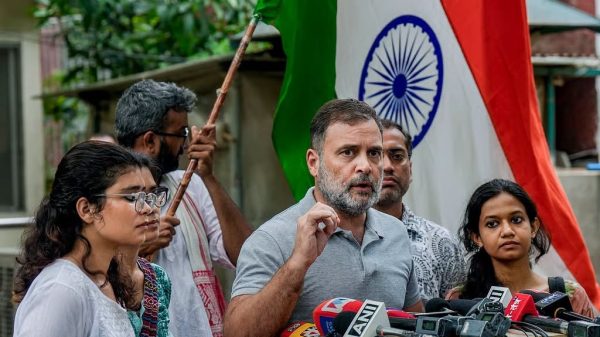 Man with gray beard addresses a crowd at a press conference, microphones in front, Indian flag in the background.