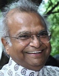 Smiling man with gray hair and glasses, wearing a patterned shirt, outdoors among green trees.