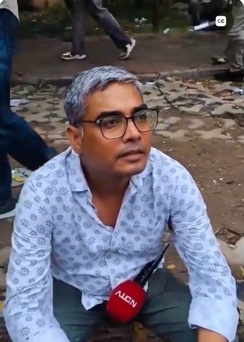 Screenshot | No. 1 Indian Media News Portal Man with gray hair and glasses sits on the ground outdoors during an interview, a red news microphone resting near his chest.