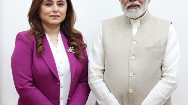 Woman in a magenta suit standing beside a man in a beige traditional-style vest, both in formal attire against a plain background.