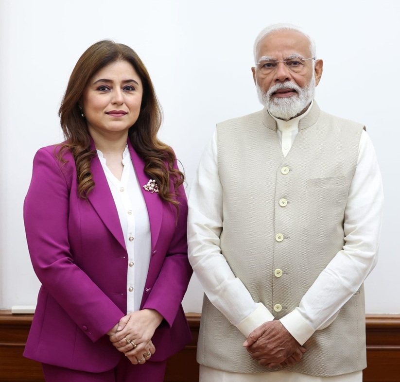 Woman in a magenta suit standing beside a man in a beige traditional-style vest, both in formal attire against a plain background.