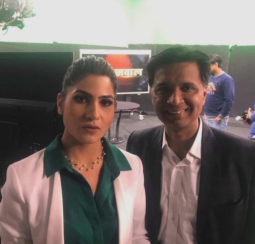 Two professionals posing for a photo in a studio; woman in a green blouse with a white blazer and a necklace, man in a dark suit and white shirt smiling next to her.