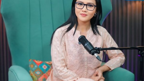 Woman with glasses sits in a teal high-back chair in a studio, speaking into a microphone on a boom arm.