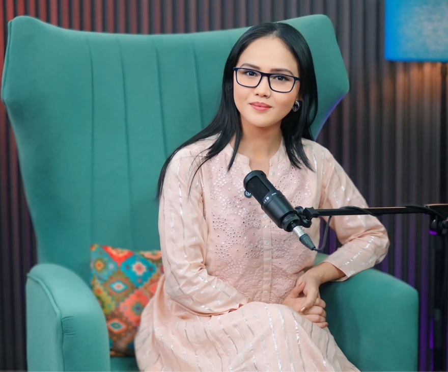 Woman with glasses sits in a teal high-back chair in a studio, speaking into a microphone on a boom arm.