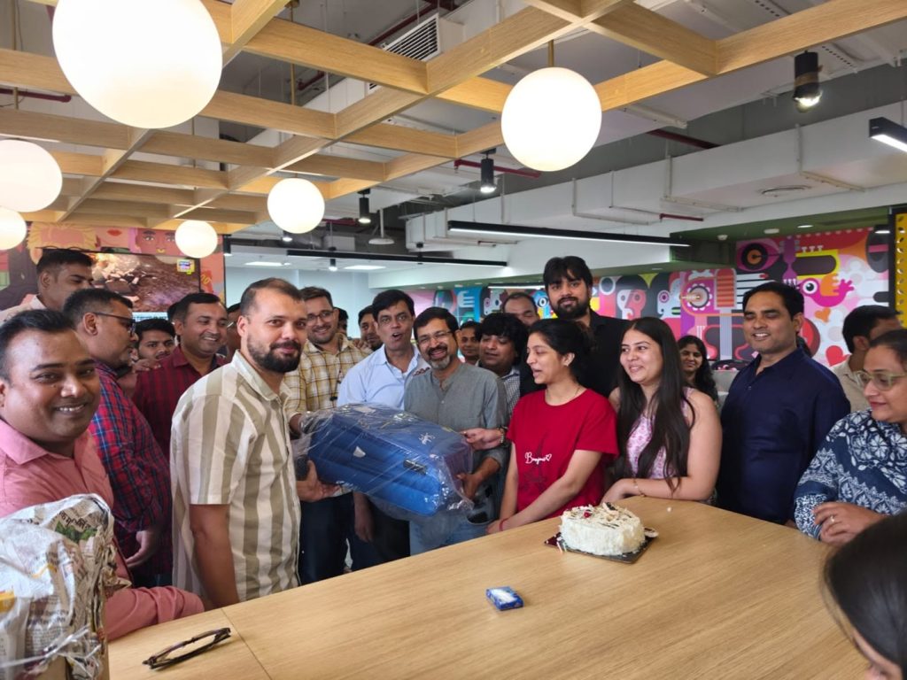 sanjay | No. 1 Indian Media News Portal Group of coworkers at a celebratory office party around a long wooden table; a man hands a large blue wrapped gift to another man, with cake on the table and colorful wall art in the background.