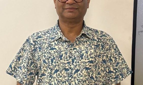 Man wearing a blue floral short-sleeve shirt and glasses, standing against a light wall and smiling slightly.