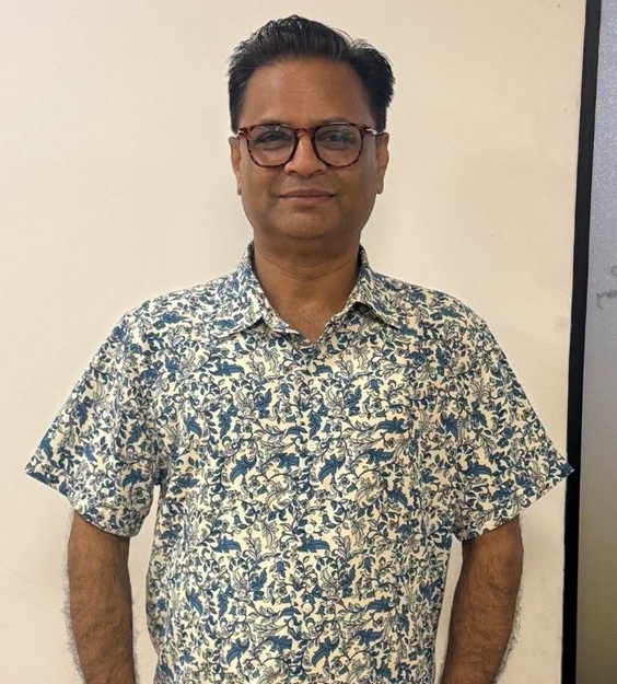 Man wearing a blue floral short-sleeve shirt and glasses, standing against a light wall and smiling slightly.