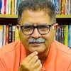 T thoughtful man wearing glasses and an orange polo sits in front of a bookshelf full of colorful books, hand on chin.