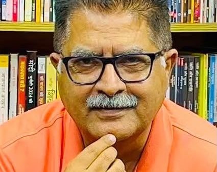 T thoughtful man wearing glasses and an orange polo sits in front of a bookshelf full of colorful books, hand on chin.
