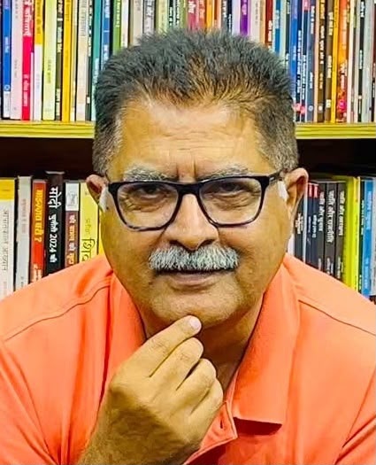 T thoughtful man wearing glasses and an orange polo sits in front of a bookshelf full of colorful books, hand on chin.