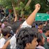 Crowd of protesters with a raised fist, an Indian flag visible among marchers in a tree-lined street with a green street sign in the background.
