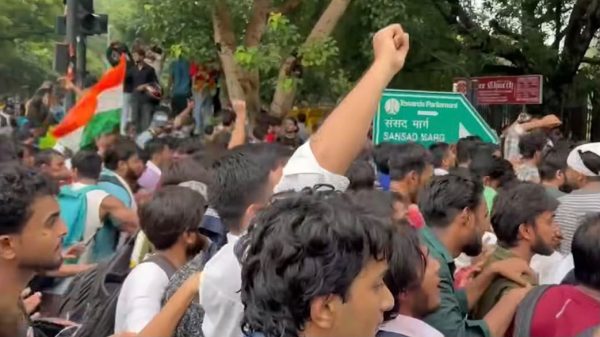 Crowd of protesters with a raised fist, an Indian flag visible among marchers in a tree-lined street with a green street sign in the background.