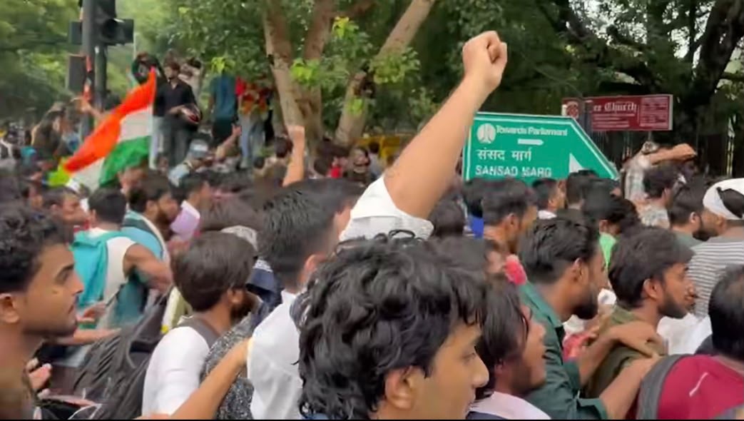 Crowd of protesters with a raised fist, an Indian flag visible among marchers in a tree-lined street with a green street sign in the background.