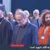 Group of dignitaries on stage at a formal event; a bearded cleric in black robes and cap stands left, others in suits and lanyards nearby.