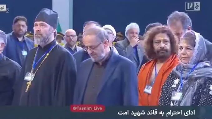 Group of dignitaries on stage at a formal event; a bearded cleric in black robes and cap stands left, others in suits and lanyards nearby.