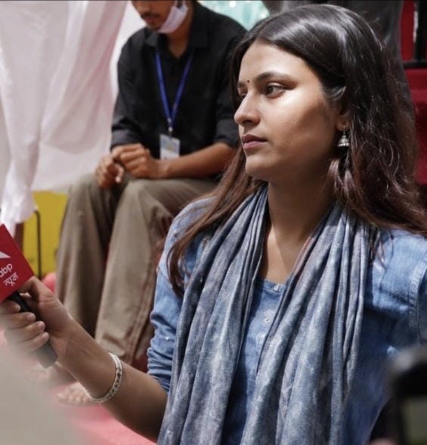 Side-profile of a woman in a blue scarf and denim top speaking into a handheld microphone during an interview