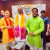Three men in traditional Indian outfits pose in a BJP office, with the party symbol on the wall behind them and colorful scarves/tilaks on their foreheads.