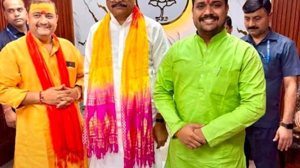 Three men in traditional Indian outfits pose in a BJP office, with the party symbol on the wall behind them and colorful scarves/tilaks on their foreheads.