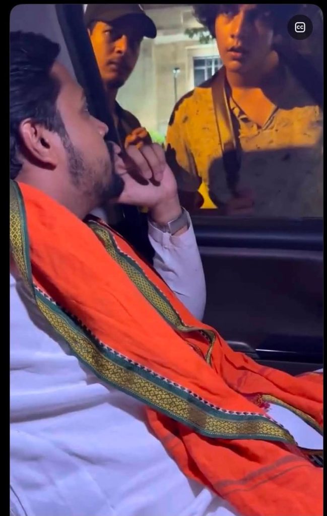 Man with a beard wearing an orange shawl sits in a car, talking to two men outside the window at night.