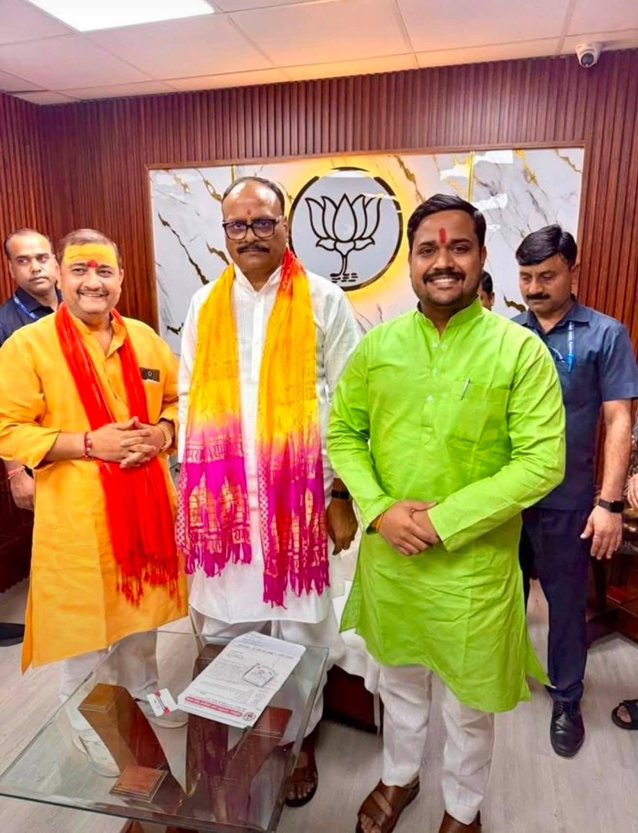 Three men in traditional Indian outfits pose in a BJP office, with the party symbol on the wall behind them and colorful scarves/tilaks on their foreheads.