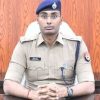 Male police officer in khaki uniform with a blue beret, seated behind a desk with hands clasped.