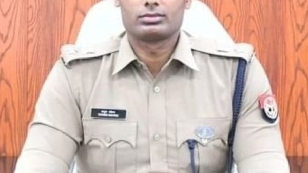Male police officer in khaki uniform with a blue beret, seated behind a desk with hands clasped.