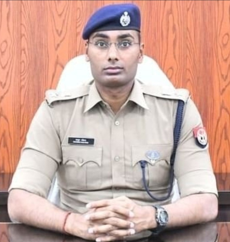 Male police officer in khaki uniform with a blue beret, seated behind a desk with hands clasped.