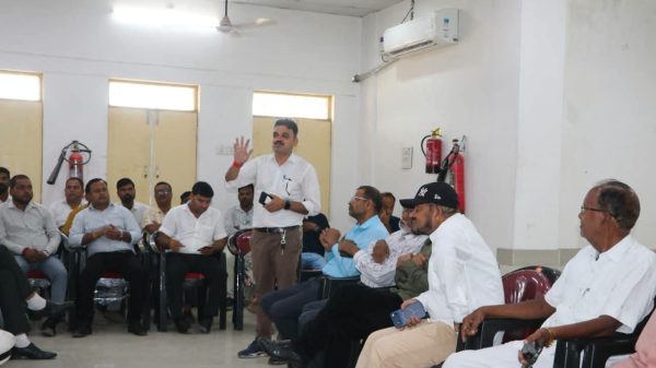 A man stands and speaks to a seated audience in a room, gesturing with one hand while others listen attentively, suggesting a workshop or meeting.