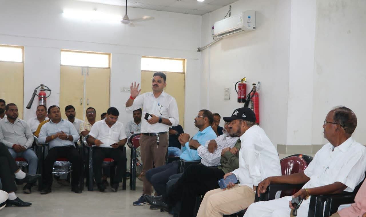 A man stands and speaks to a seated audience in a room, gesturing with one hand while others listen attentively, suggesting a workshop or meeting.