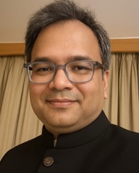 shailendra jha | No. 1 Indian Media News Portal Portrait of a man with gray hair and glasses, wearing a black high-collar jacket, smiling in front of beige curtains.