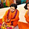 Two men in orange robes sit on a white cloth, performing a devotional ritual with a plate of fruit and marigold flowers.