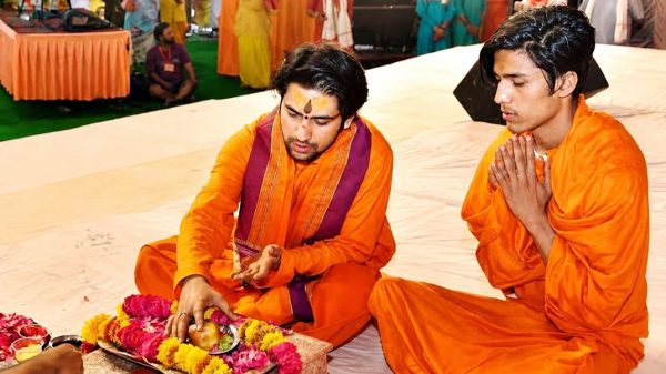 Two men in orange robes sit on a white cloth, performing a devotional ritual with a plate of fruit and marigold flowers.