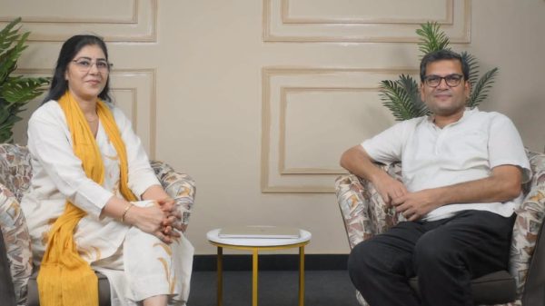 Two adults sit in patterned armchairs in a living room, with a small round yellow-and-white table between them and a closed laptop on the table.