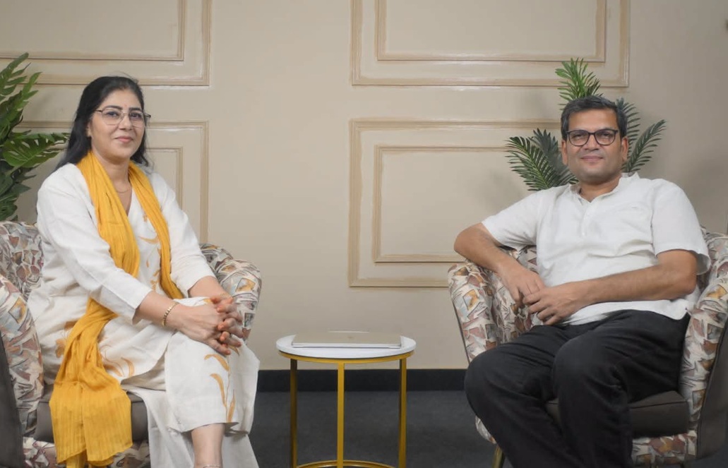 Two adults sit in patterned armchairs in a living room, with a small round yellow-and-white table between them and a closed laptop on the table.