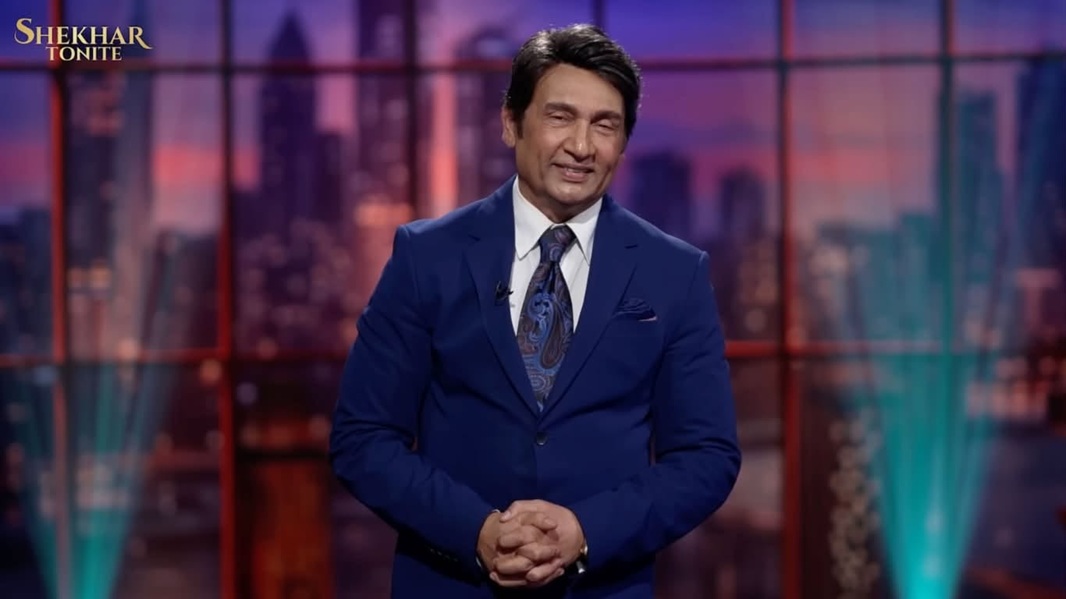 Male host in a blue suit on the Shekhar Tonight TV set, smiling with hands clasped in front of a cityscape backdrop.