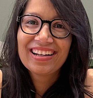 Close-up portrait of a smiling woman with shoulder-length dark hair and black-framed glasses facing the camera.
