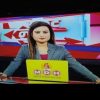 Female news anchor sitting at a news desk with a yellow laptop, red studio backdrop behind her.