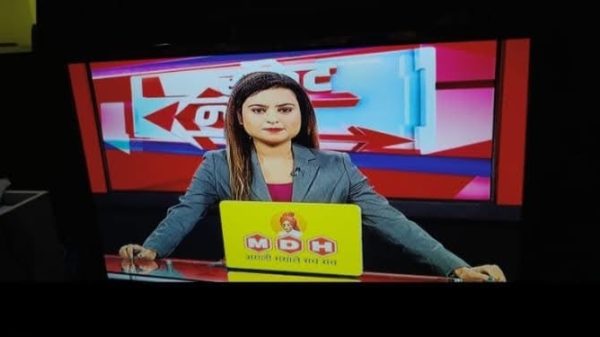 Female news anchor sitting at a news desk with a yellow laptop, red studio backdrop behind her.