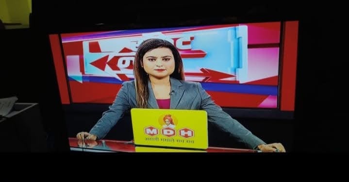 Female news anchor sitting at a news desk with a yellow laptop, red studio backdrop behind her.