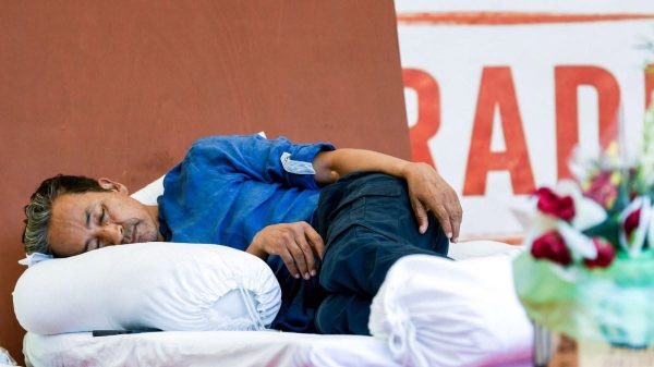 Man sleeping on a white pillow on a bed, wearing a blue shirt, with flowers in the foreground and red lettering in the background.