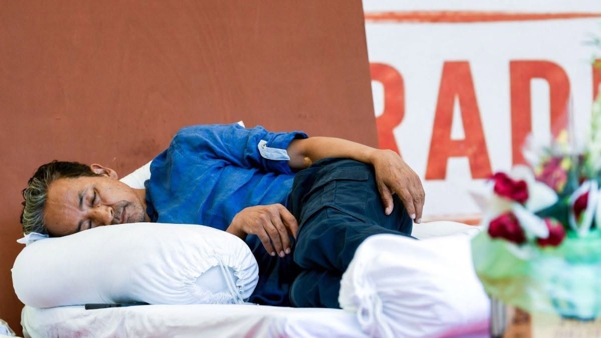 Man sleeping on a white pillow on a bed, wearing a blue shirt, with flowers in the foreground and red lettering in the background.