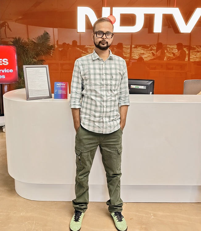 oplus_134217760 | No. 1 Indian Media News Portal Man in a light plaid shirt and olive cargo pants stands with hands in pockets at a white reception desk in front of a bright orange NDTV sign.