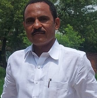 Man in a white button-down shirt with a pen in the chest pocket, standing outdoors with greenery in the background.